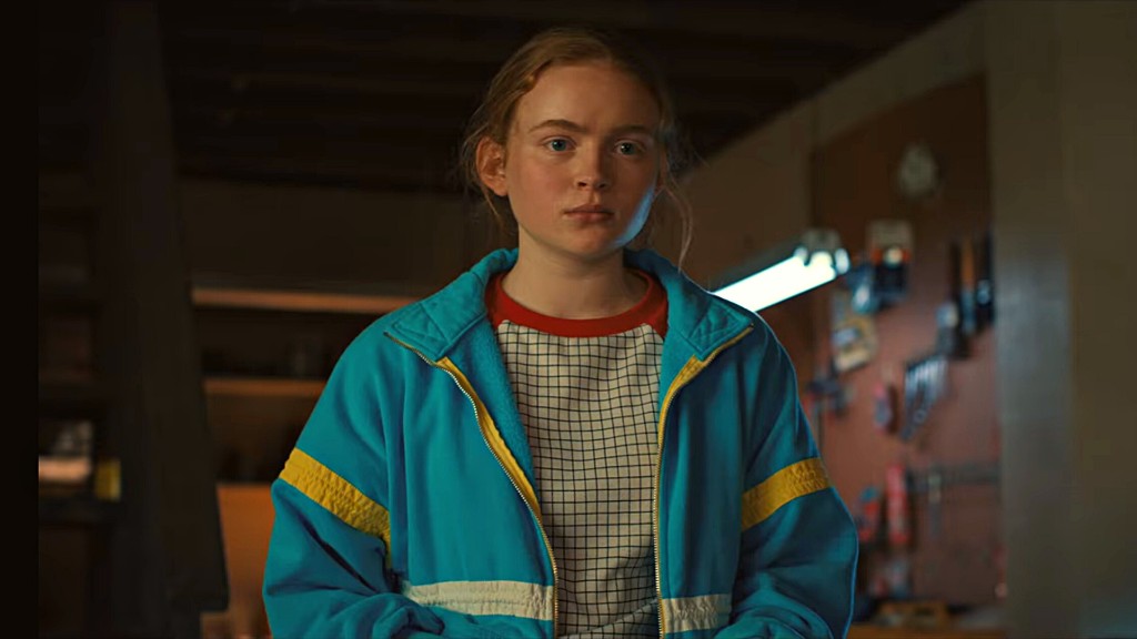 Max Mayfield in een blauw jack met gele strepen en rode kraag, in een werkplaats