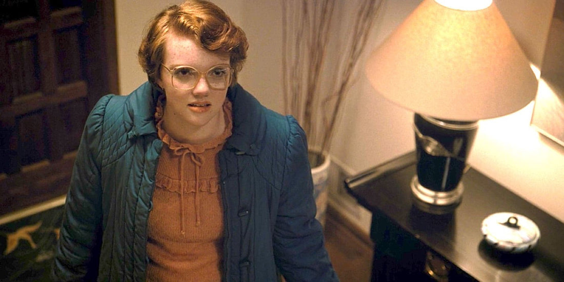 Shannon Purser als Barbara Holland, bril en blauwe jas, in een hal.